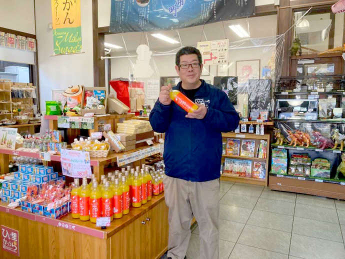 物産館の店内で勝浦みかんストレートを持ってほほ笑む渡邊さん