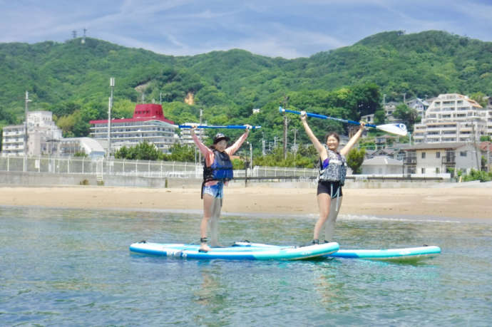 穏やかな浅瀬の海岸でボードの上に立ちパドルを掲げる女性