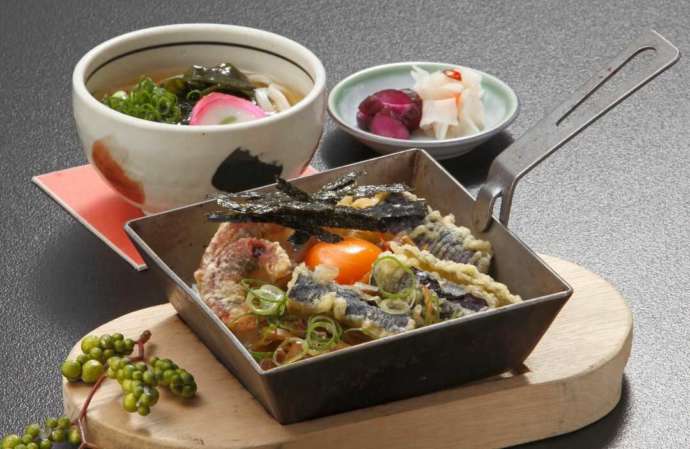 正方形の鉄鍋に入って卵黄の乗った天丼と、うどん、漬物のセット